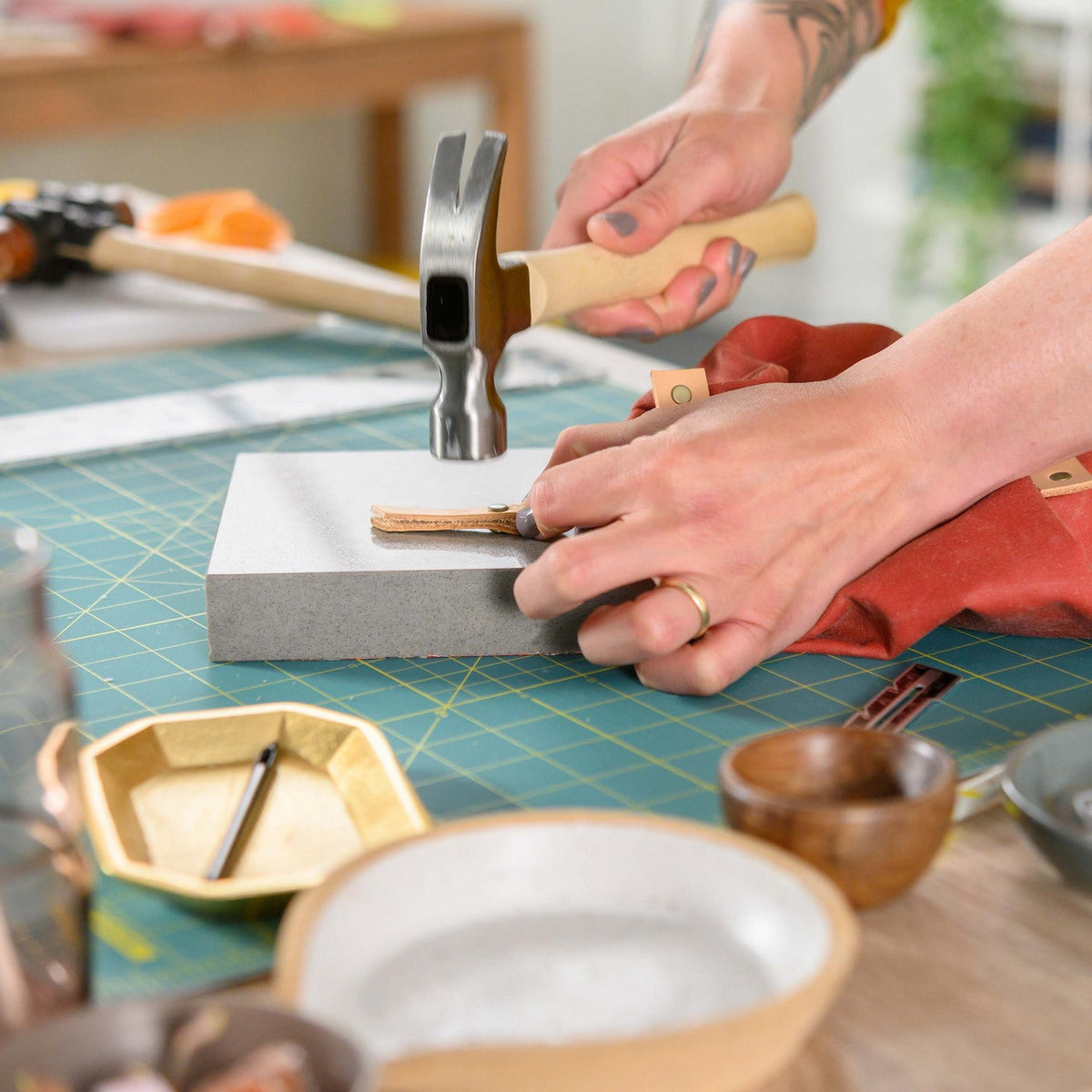 A maker holding a leather zipper pull flat on a granite slab and setting its rivet with a hammer, beside a rust waxed canvas bag on a teal cutting mat.