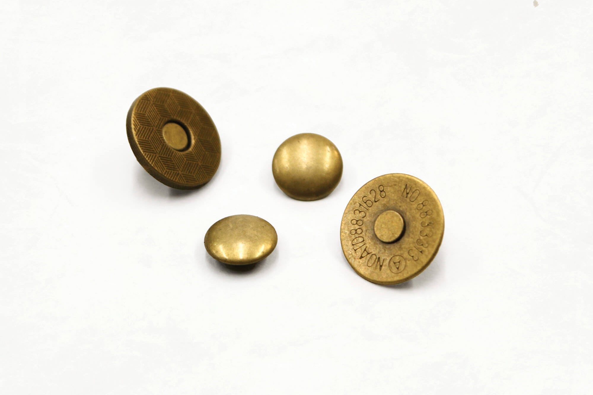 The four pieces of a magnetic snap rivet in antique brass on a white surface: two smooth domed caps, plus the two magnet halves, one with a cross-hatched face and one stamped with its size code.