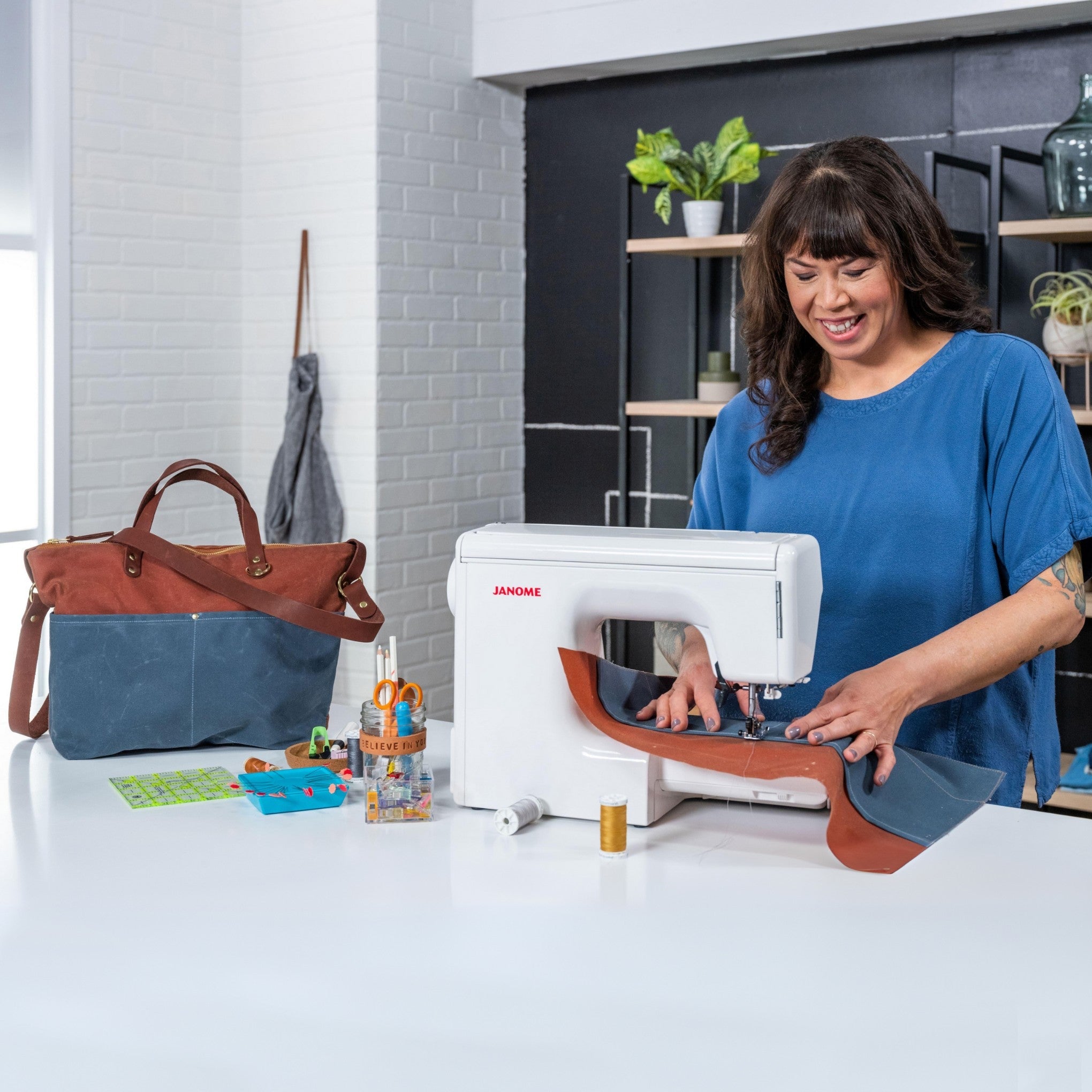 A maker sewing waxed canvas on a white Janome home sewing machine, with a finished blue and brown tote bag beside her