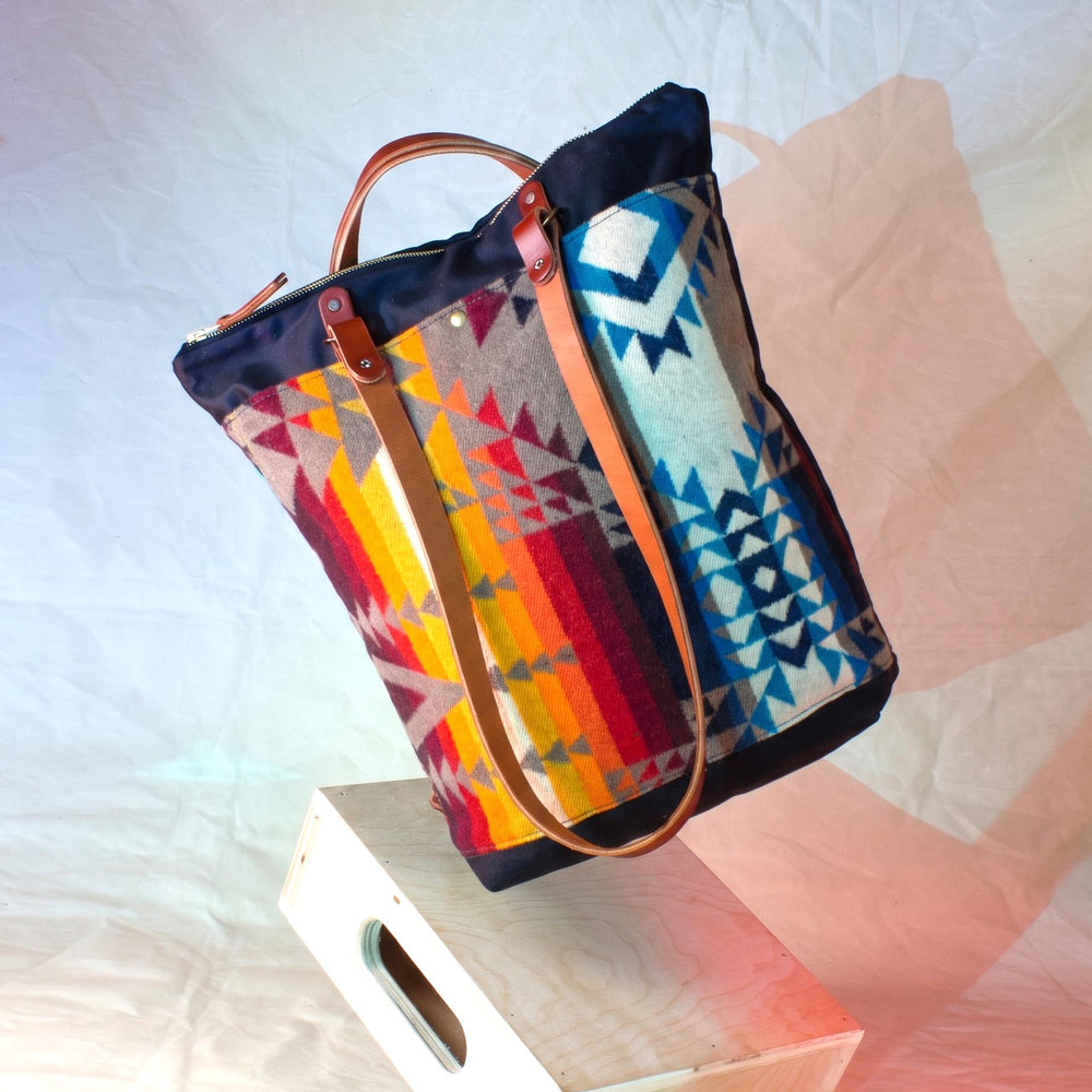 A woven tote bag with a Southwestern geometric pattern in red, orange, yellow, blue, and gray, featuring tan leather handles and straps, resting on a white wooden block against a white fabric backdrop.