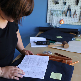 A maker at a worktable reading a Klum House instruction booklet, with navy waxed canvas, leather straps and a mallet laid out and a pegboard of tools behind her.