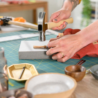 A maker holding a leather zipper pull flat on a granite slab and setting its rivet with a hammer, beside a rust waxed canvas bag on a teal cutting mat.