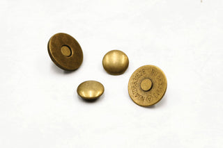 The four pieces of a magnetic snap rivet in antique brass on a white surface: two smooth domed caps, plus the two magnet halves, one with a cross-hatched face and one stamped with its size code.
