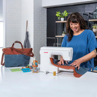 A maker sewing waxed canvas on a white Janome home sewing machine, with a finished blue and brown tote bag beside her