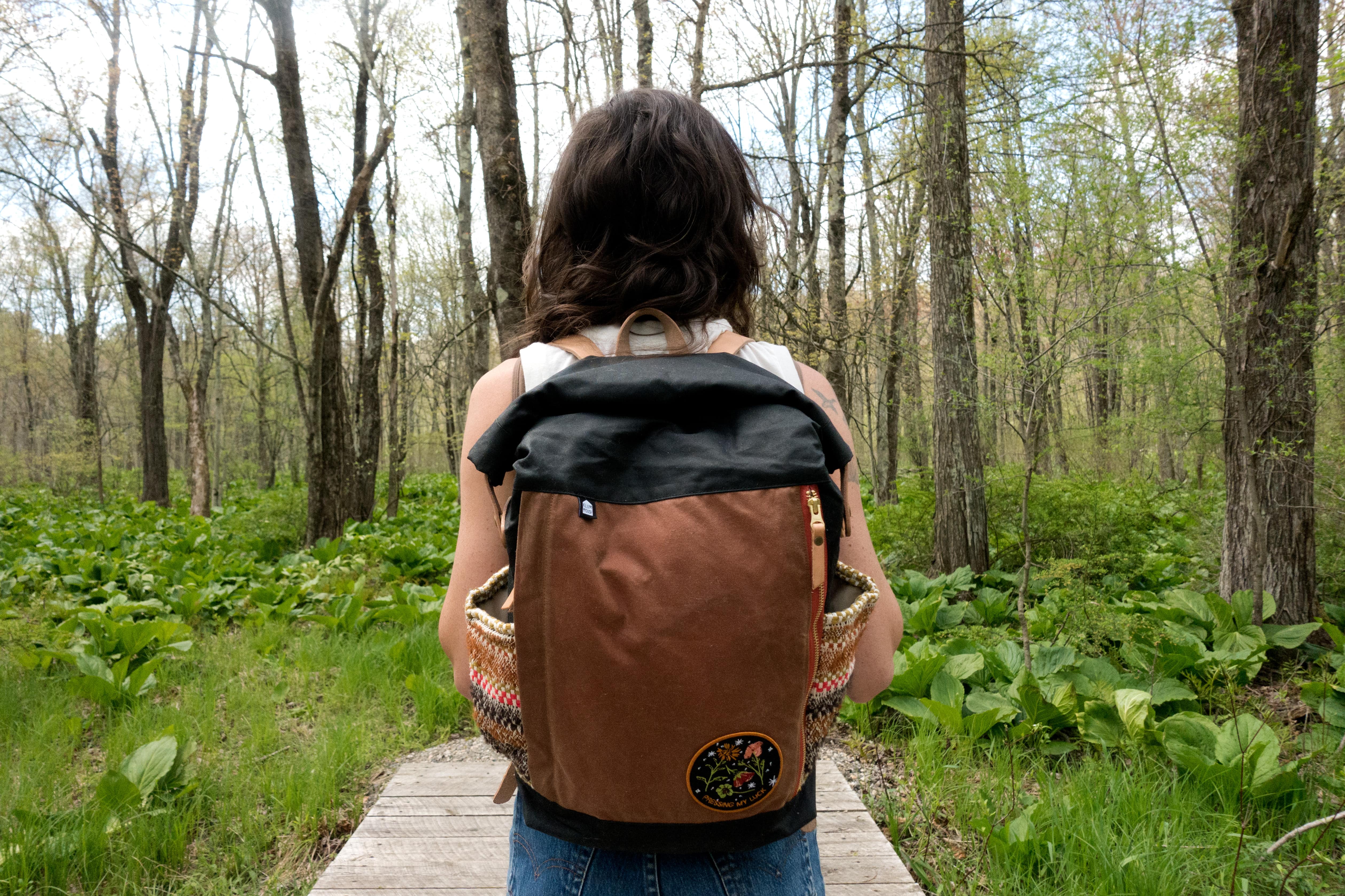 Katie Whittle on a forest boardwalk, back to camera, wearing her finished Slabtown Backpack with its patterned woven side pockets and embroidered patch