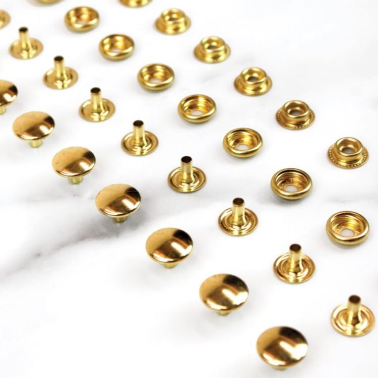 A scatter of brass Line 24 snap pieces on white marble: domed caps with posts standing up, and sockets showing the ring that receives the stud.