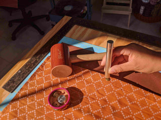 A hand positions a metal punch over patterned orange fabric, next to a Bakelite mallet, a metal ruler and a dish of rivets.